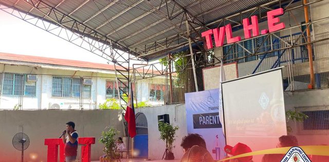 A speaker on the campus stage under the TVL H.E sign during a parents' meeting.